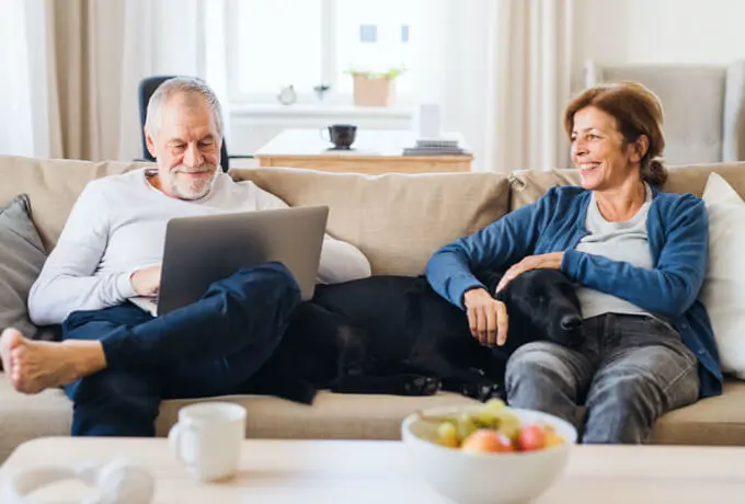 Older couple sitting on couch and man is looking at laptop. Bowl of apples sit on table in front of them