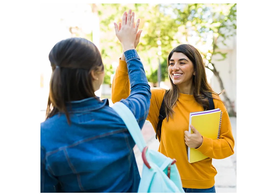 Female student carrying her books and notebooks while giving high fives to her college friend